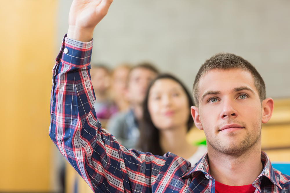 Close-up of a young male student raising hand by others in a row at the classroom